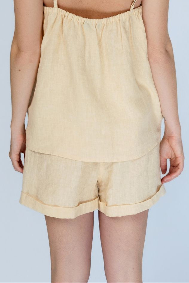 A woman shown from the back wearing a light beige linen romper with thin spaghetti straps and matching shorts, set against a solid light blue background.