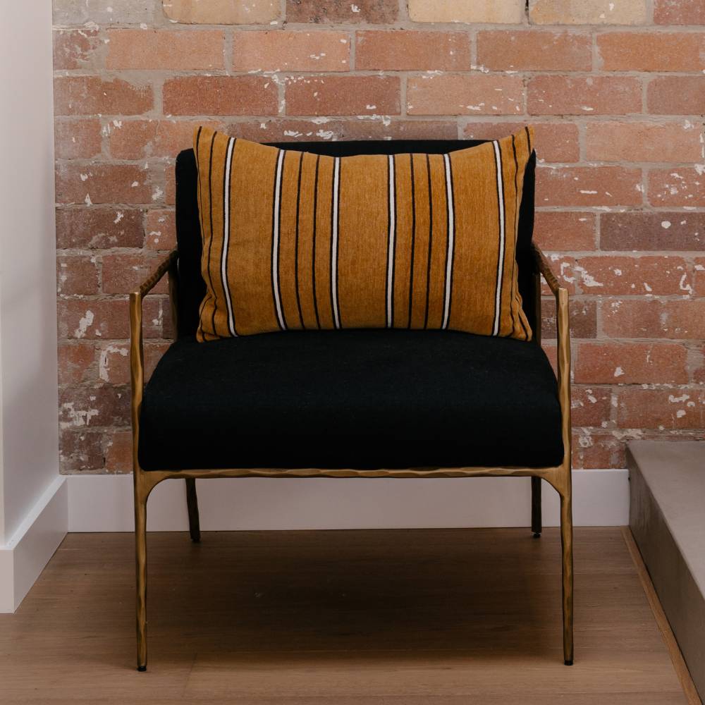 Brown striped pillow on a chair with black seat and gold frame against a brick wall