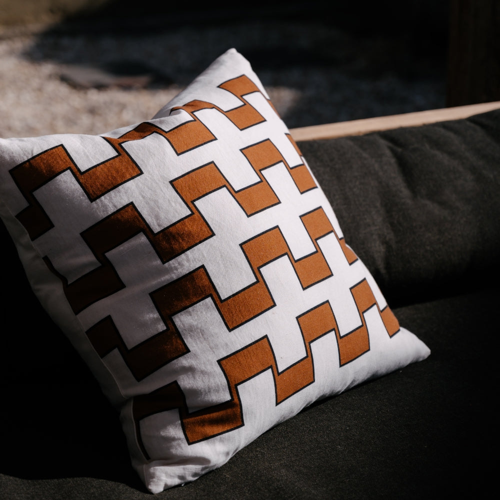 A square pillow with an interlocking brown and white geometric pattern, outlined in black, rests on a dark gray cushion.