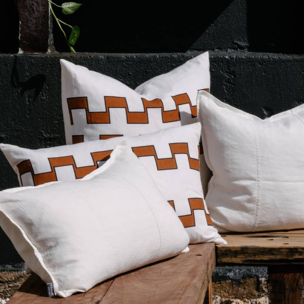 Decorative pillows on a wooden bench against a dark brick wall. Two pillows have an interlocking brown and white geometric pattern.