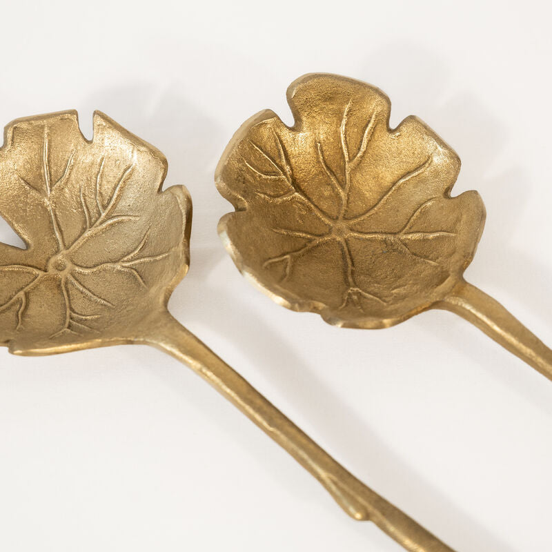 Close-up look of a set of gold-colored salad servers with a leaf design on a white background
