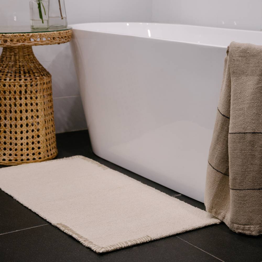 Cream or off white woven bathmat and towel on a dark tiled bathroom floor with a white bathtub.