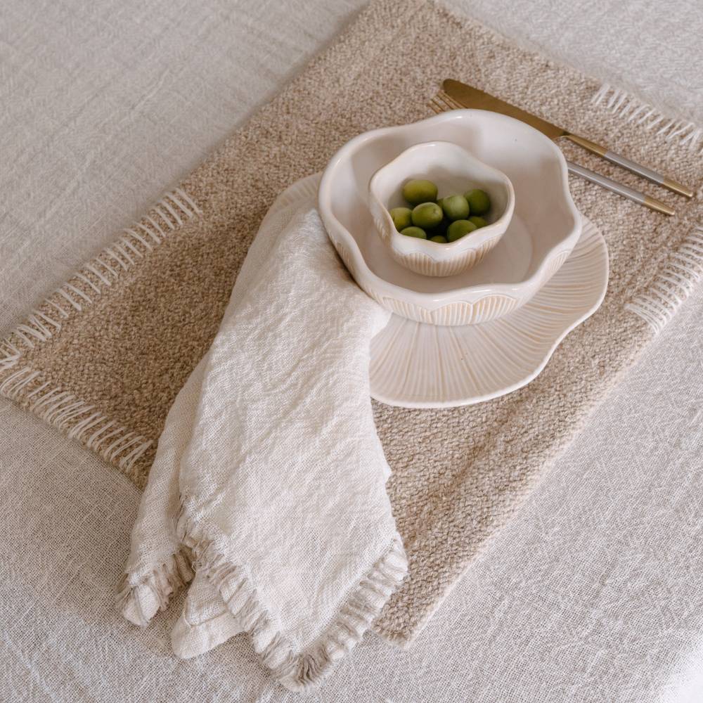 A white linen table napkin is draped over a tan placemat next to a white bowl filled with green olives sits on a larger, similarly fluted plate, next to a gold knife and fork.