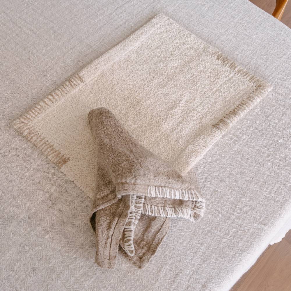 A textured cream-colored placemat with tan striped edges rests on a light-colored tablecloth, with a folded brown linen napkin placed on top of it.