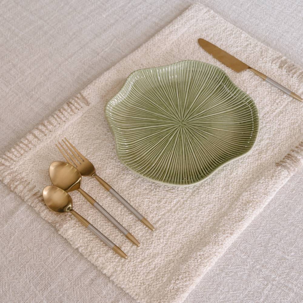 A cream placemat holds a green, fluted plate and gold utensils (two spoons, fork, knife) on a light tablecloth