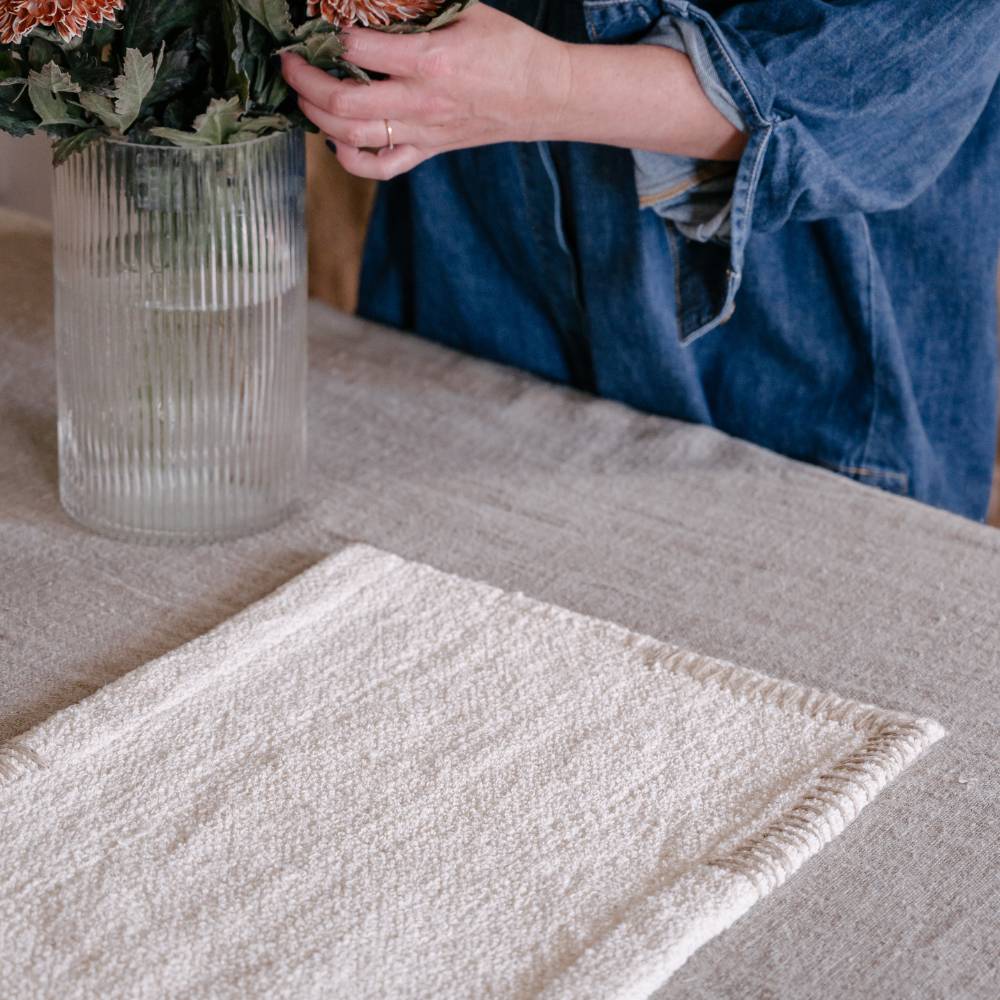 A cream-colored rectangular placemat rests on a gray linen tablecloth. In the background, a person in a denim shirt arranges flowers in a ribbed glass vase
