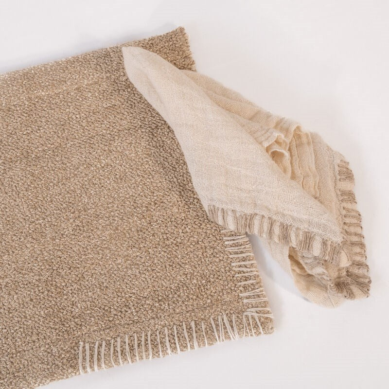 A close-up of a tan placemat with white fringe and a folded beige linen napkin with similar fringe resting on it.
