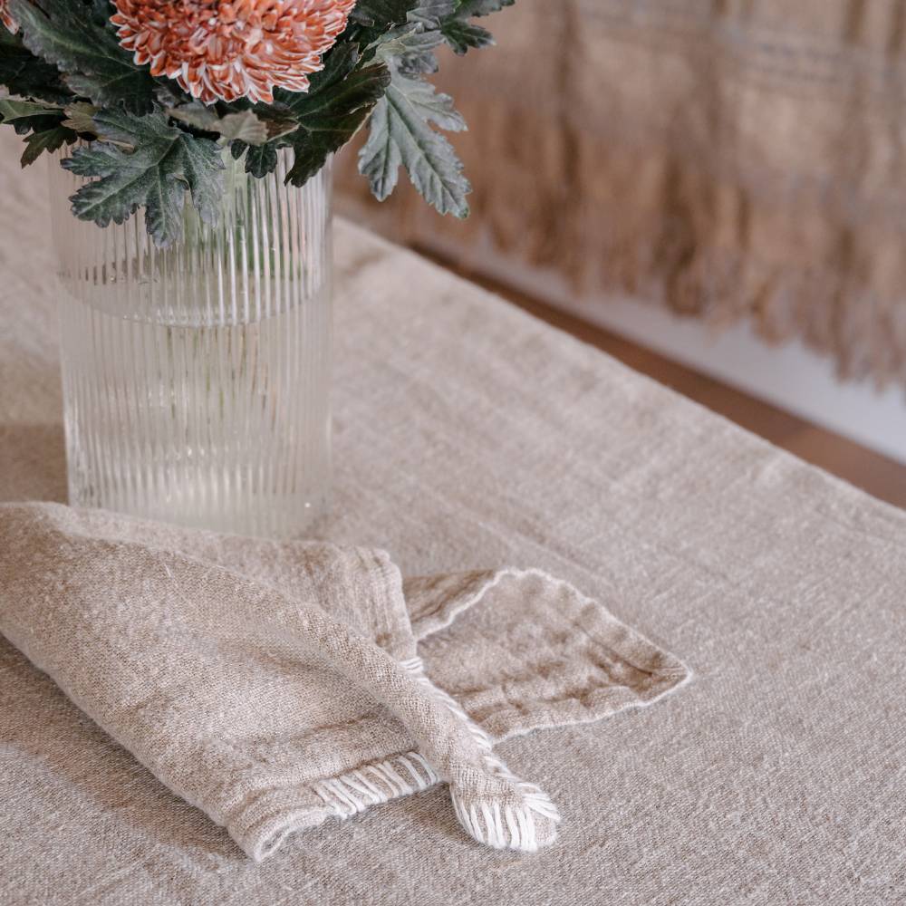 Beige table napkin on a textured natural table cloth surface with a vase of flowers in the background