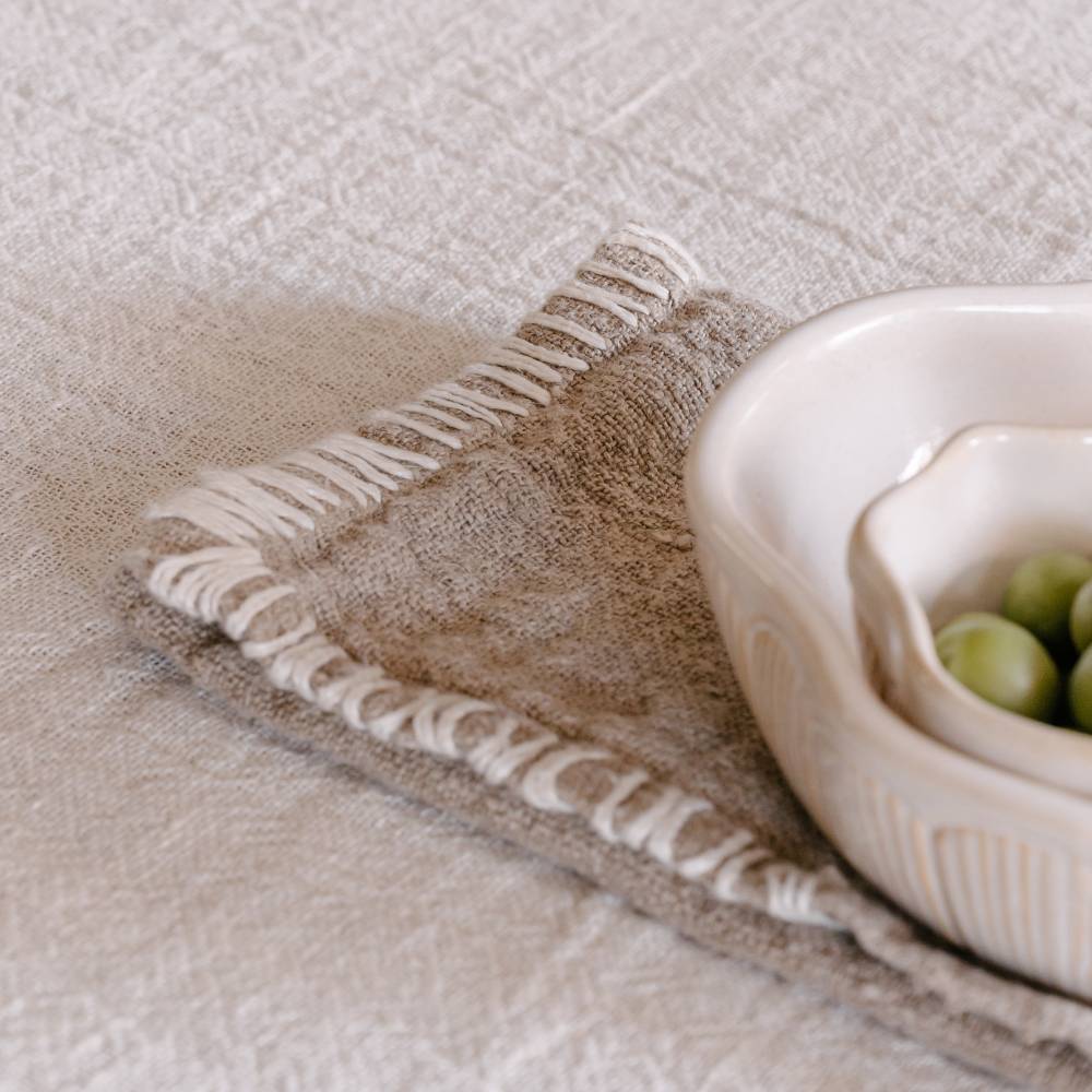 A textured beige linen table napkin with a decorative stitched edge is draped over a light tablecloth, holding two bowls.