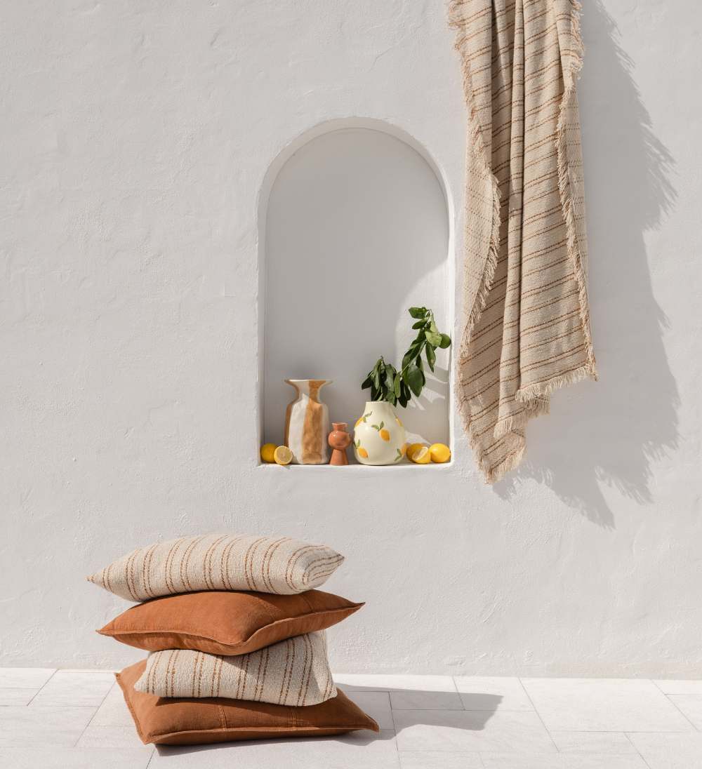 Stack of linen Eadie cushions in nutmeg and neutral  against a white wall with decorative items on a shelf.