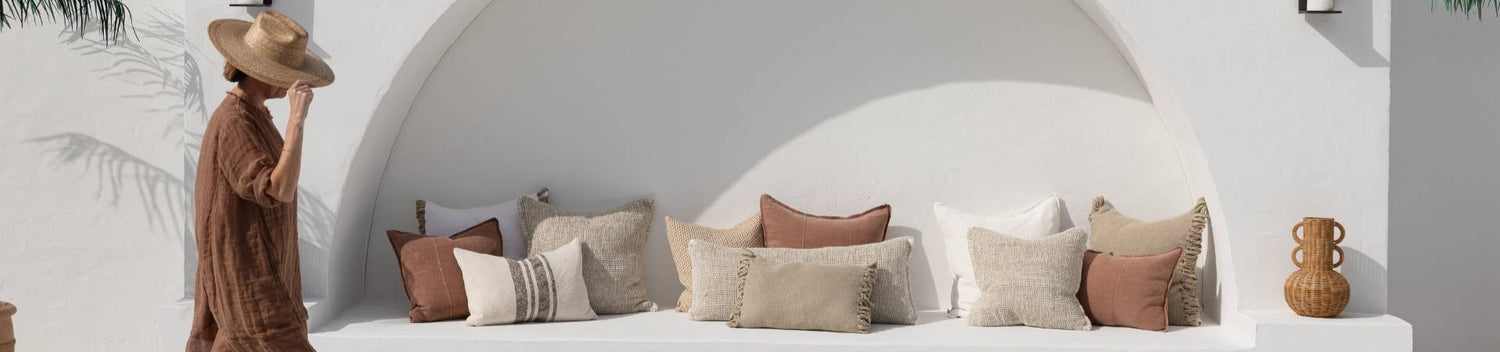 Person in a straw hat walking pass a white wall with decorative linen Eadie cushions and a vase with a Mediterranean vibe.