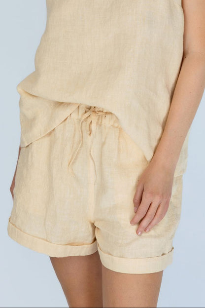 A woman wearing a light beige linen romper with a camisole-style top and matching drawstring shorts, shown against a pale blue studio background.