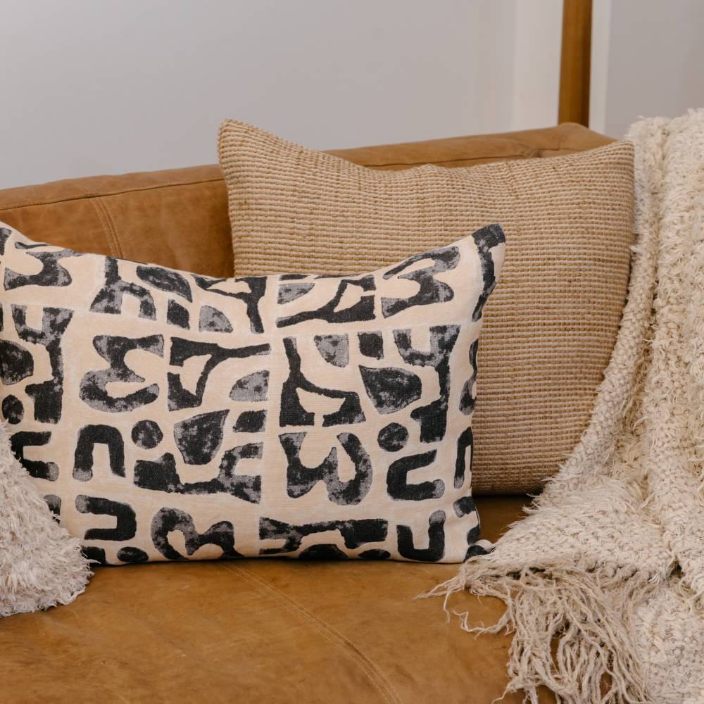 Decorative pillow with black abstract pattern on beige fabric, placed on a brown sofa.