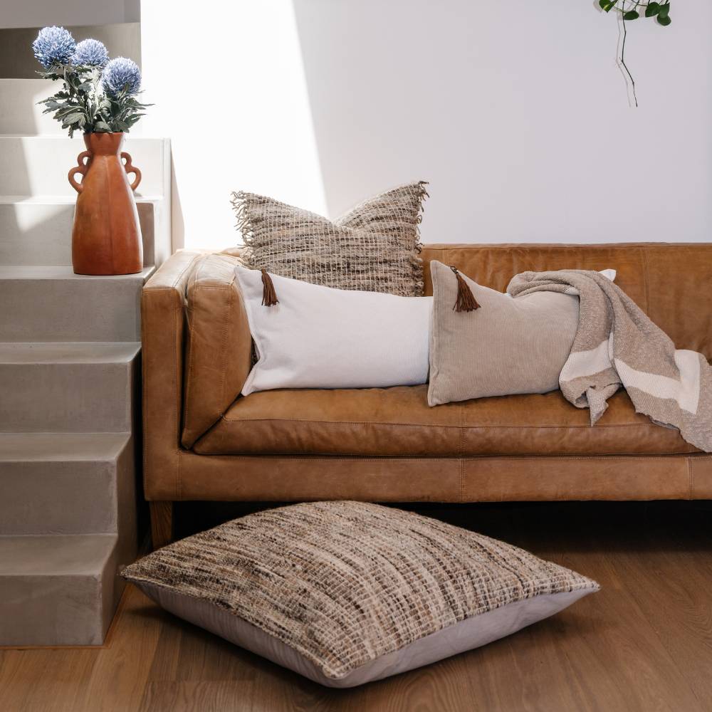 Brown sofa with patterned and plain cushions, a vase with flowers, and a plant on a white wall.
