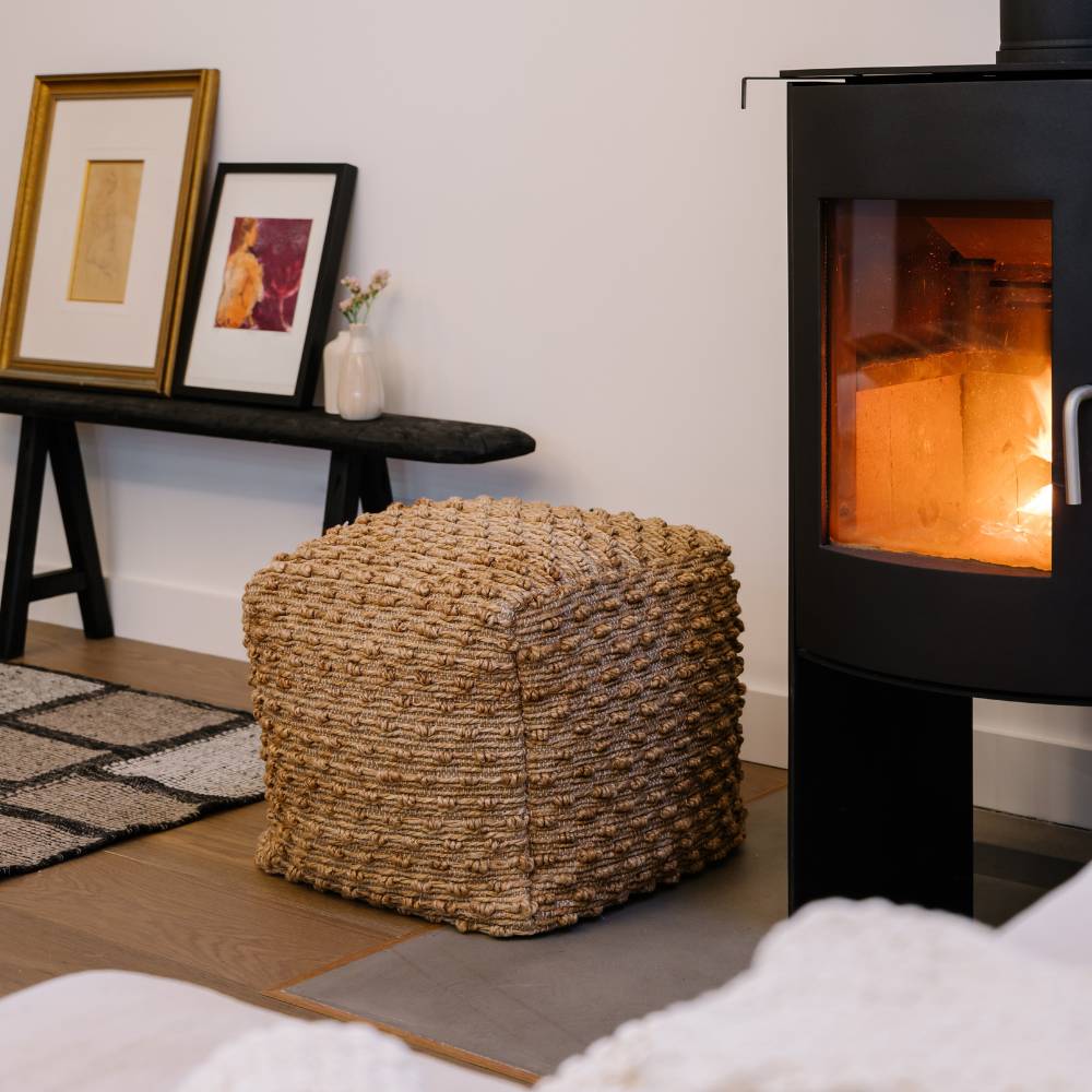 Woven ottoman in front of a lit wood stove with framed pictures on a shelf in the background.