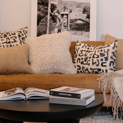 Brown sofa with patterned and fluffy pillows, books on a coffee table, and a framed picture in the background.