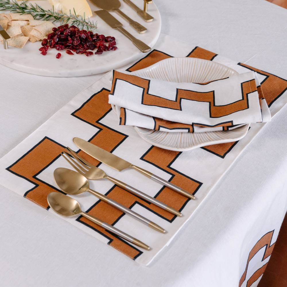 Table setting with geometric placemat, gold cutlery, and white plates on a white tablecloth.