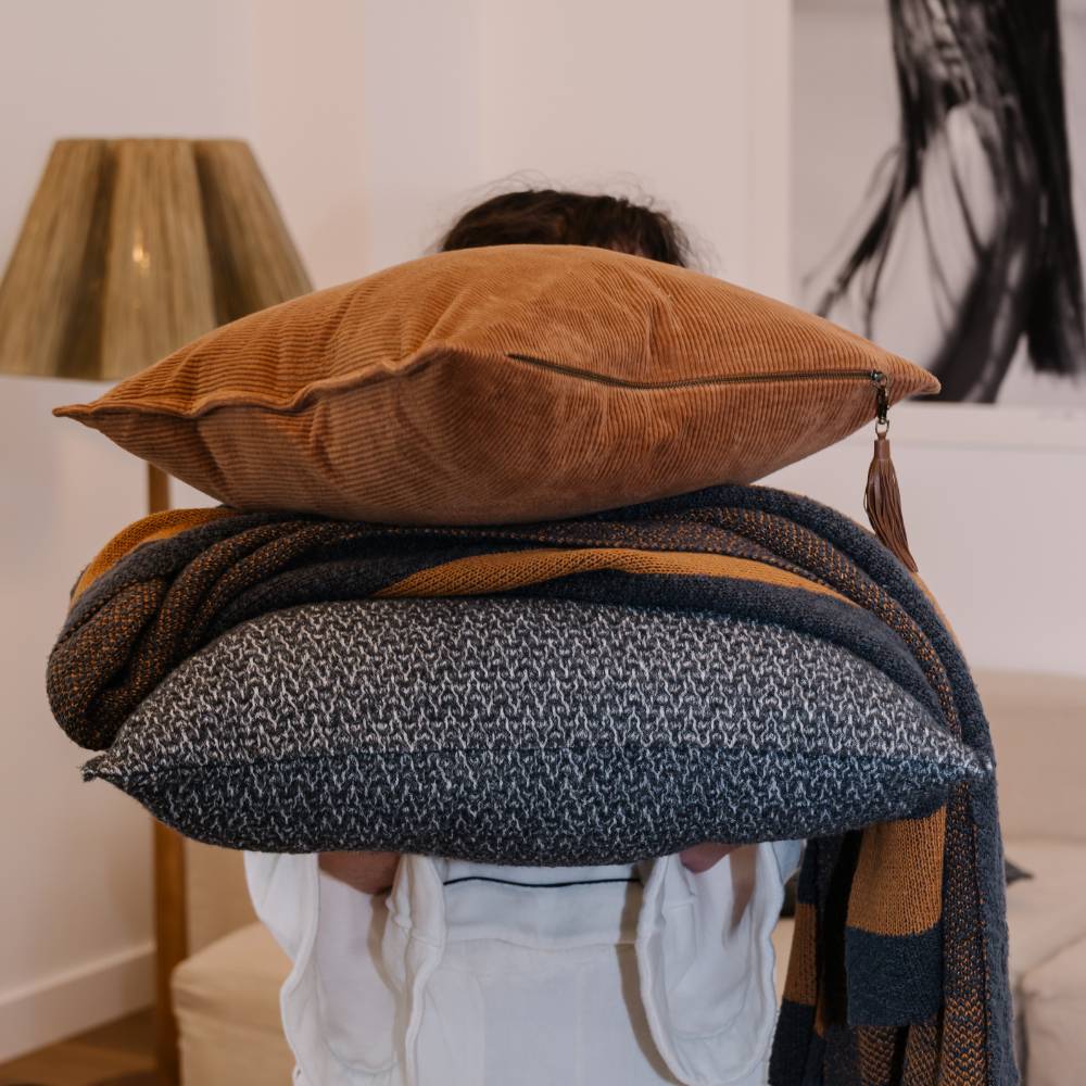 Person holding a stack of pillows and blankets in a room with a lamp and abstract painting.