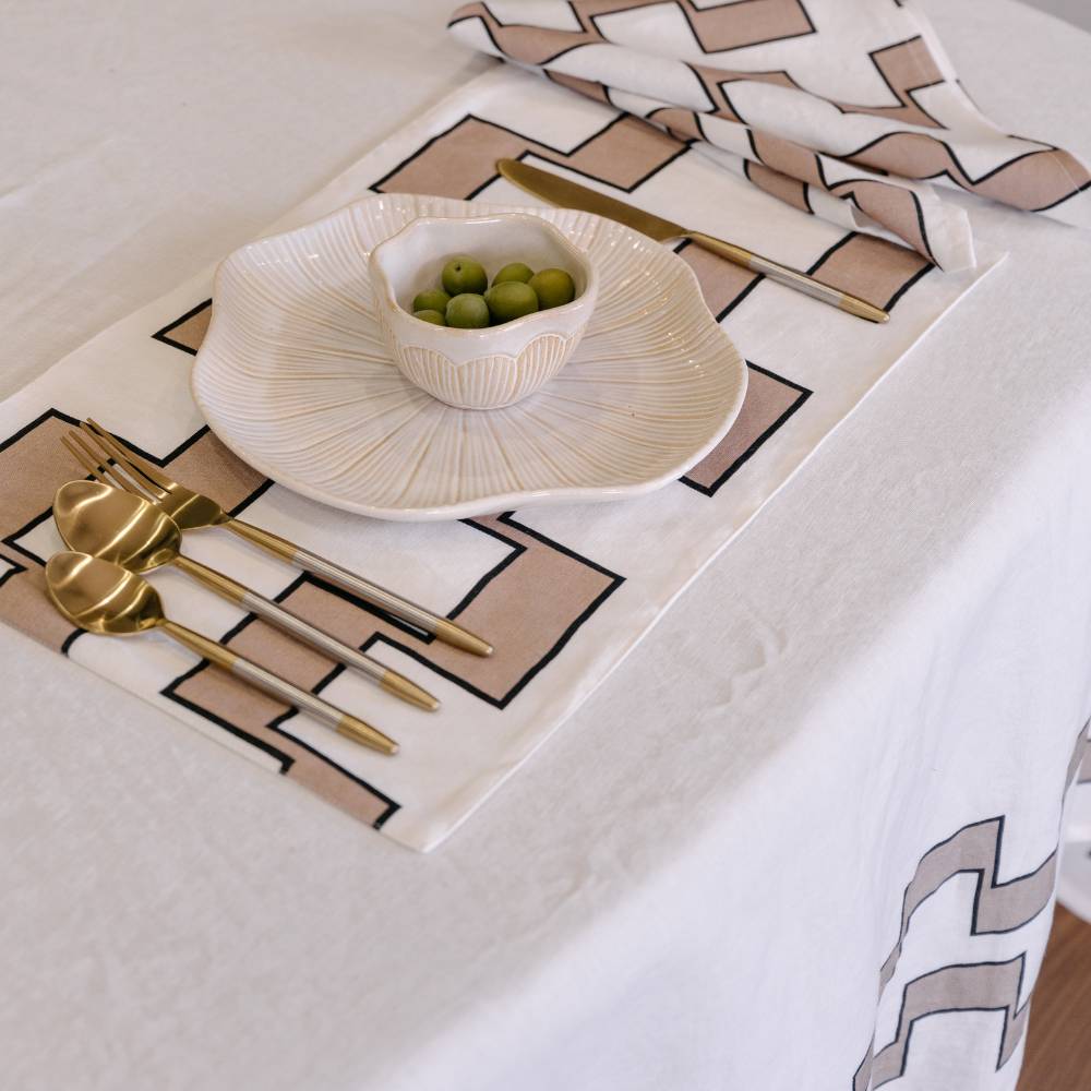 Table setting with geometric placemats, gold cutlery, and a bowl of olives on a white tablecloth.