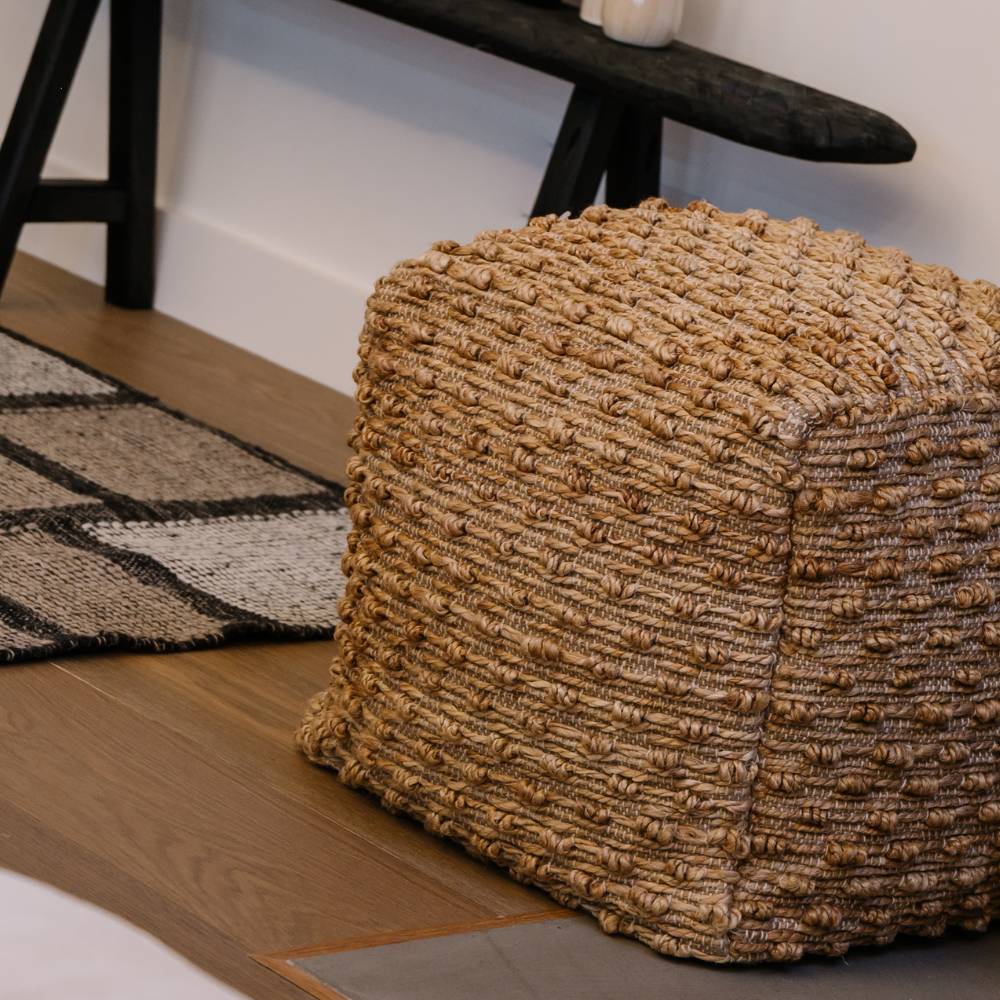 Textured woven ottoman on a wooden floor with a rug and black chair in the background.
