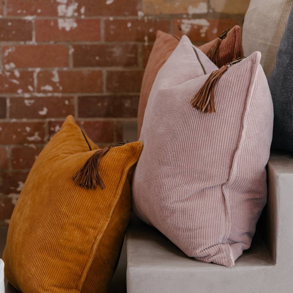 Pink and mustard yellow pillows with tassels on a white surface against a brick wall.