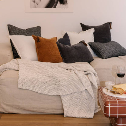 Cozy living room with a beige sofa adorned with various pillows and a blanket, accompanied by a small table with snacks and a glass of red wine.