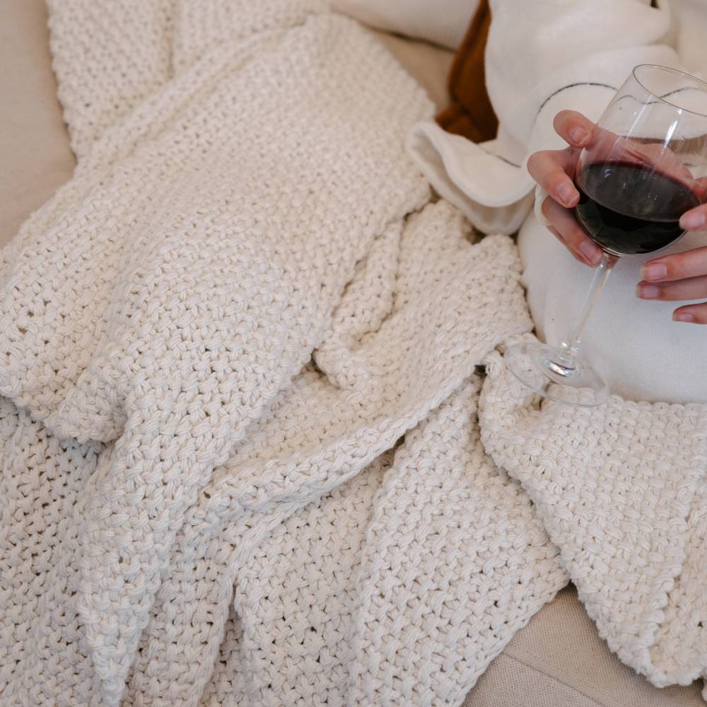 Person holding a glass of red wine on a couch with a white knit blanket