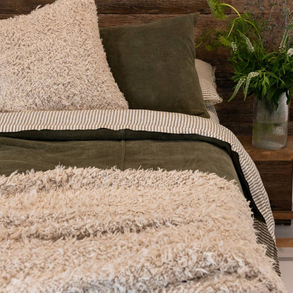 Close-up of a textured beige blanket on a bed with pillows and a plant in the background.