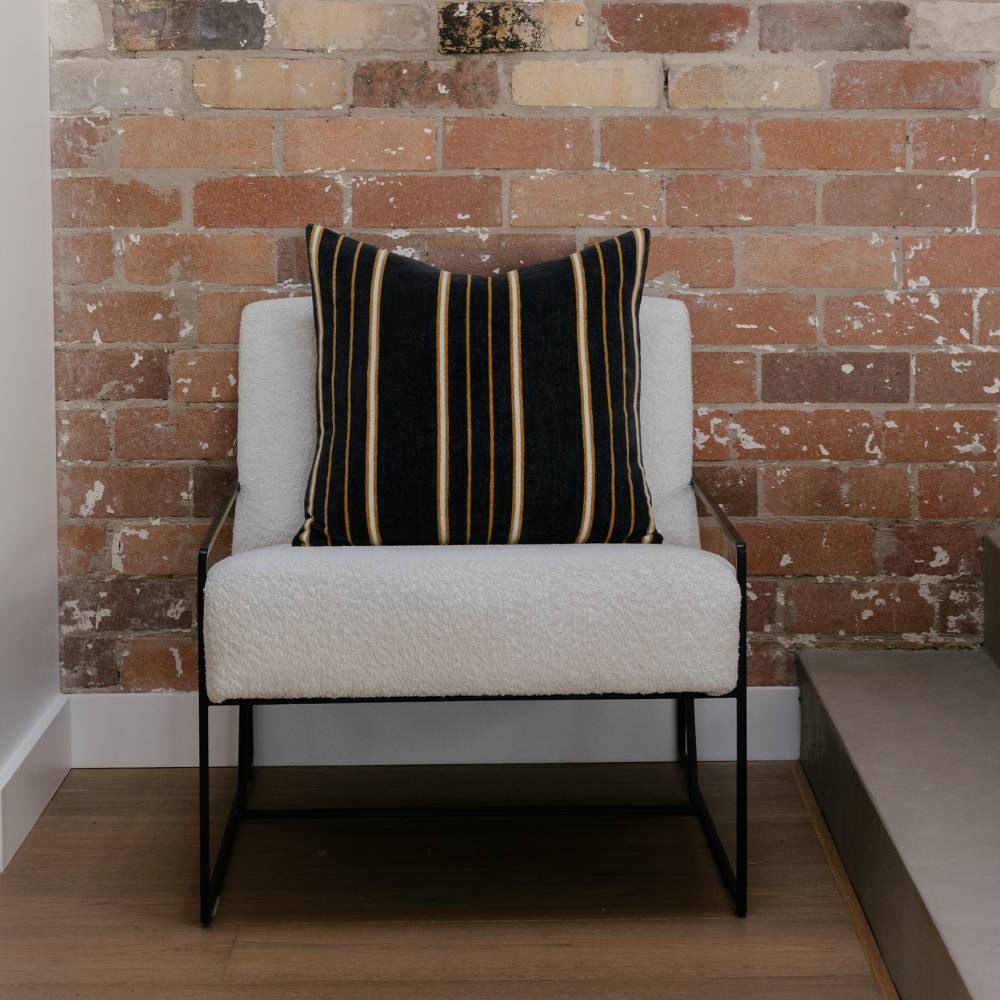 White armchair with a black, white and brown striped pillow against a brick wall.