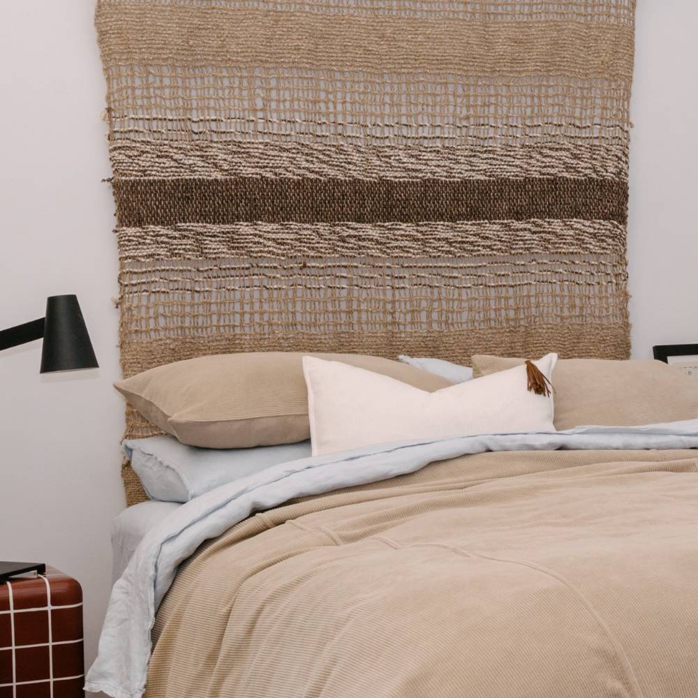 Bed with beige bedding and pillows against a textured woven headboard.