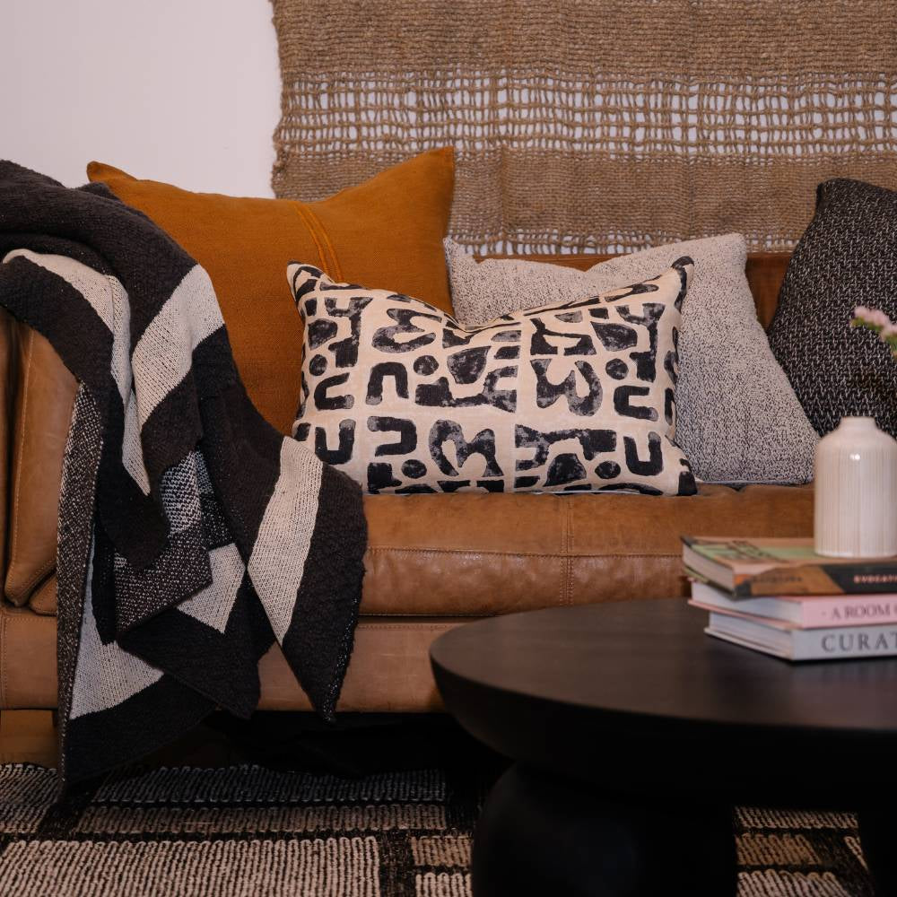 Brown leather sofa with patterned cushions and a blanket, next to a dark wooden table with books.