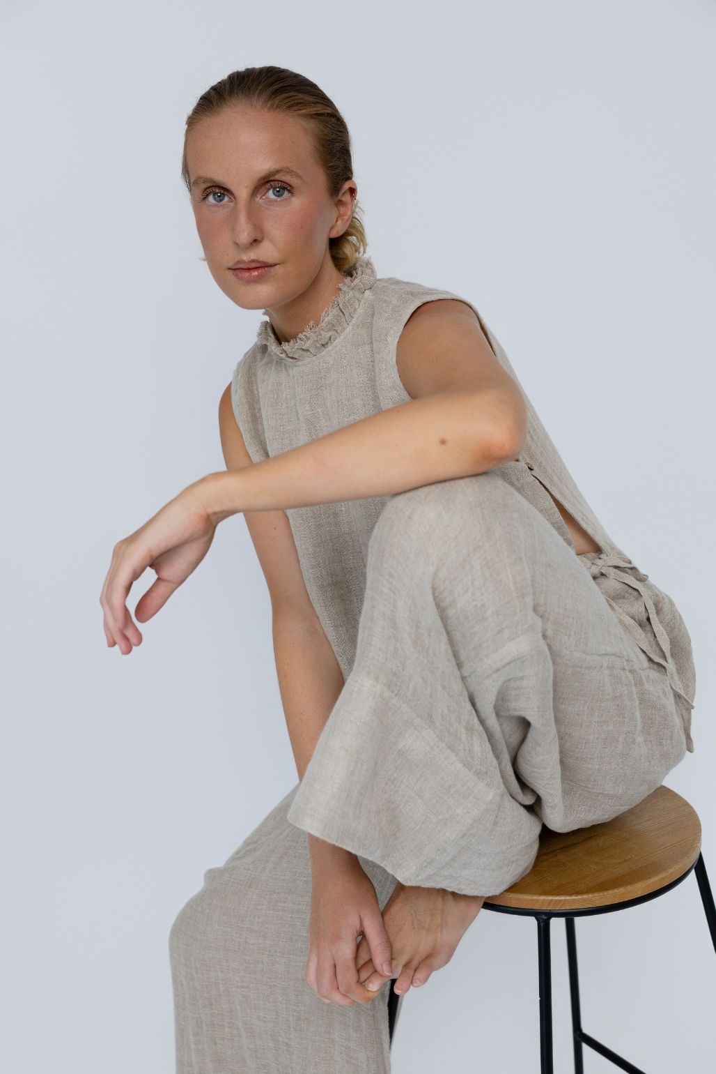 Woman sitting on a stool wearing a linen sleeveless top and pants against a plain background
