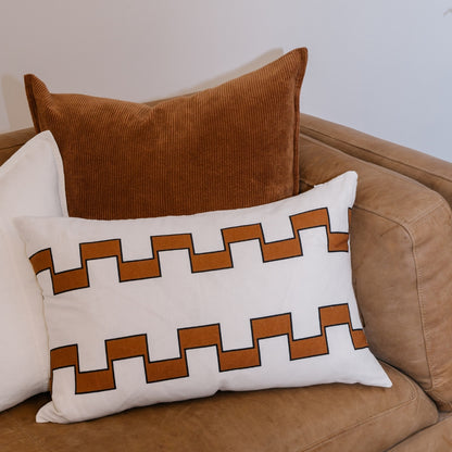 A rectangular pillow with a geometric interlocking pattern. Behind it are a solid white pillow and a ribbed brown pillow resting on a brown couch.
