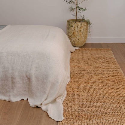 Bed with white bedding on a wooden floor next to a natural handwoven jute rug and plant