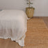 Bed with white bedding on a wooden floor next to a natural handwoven jute rug and plant