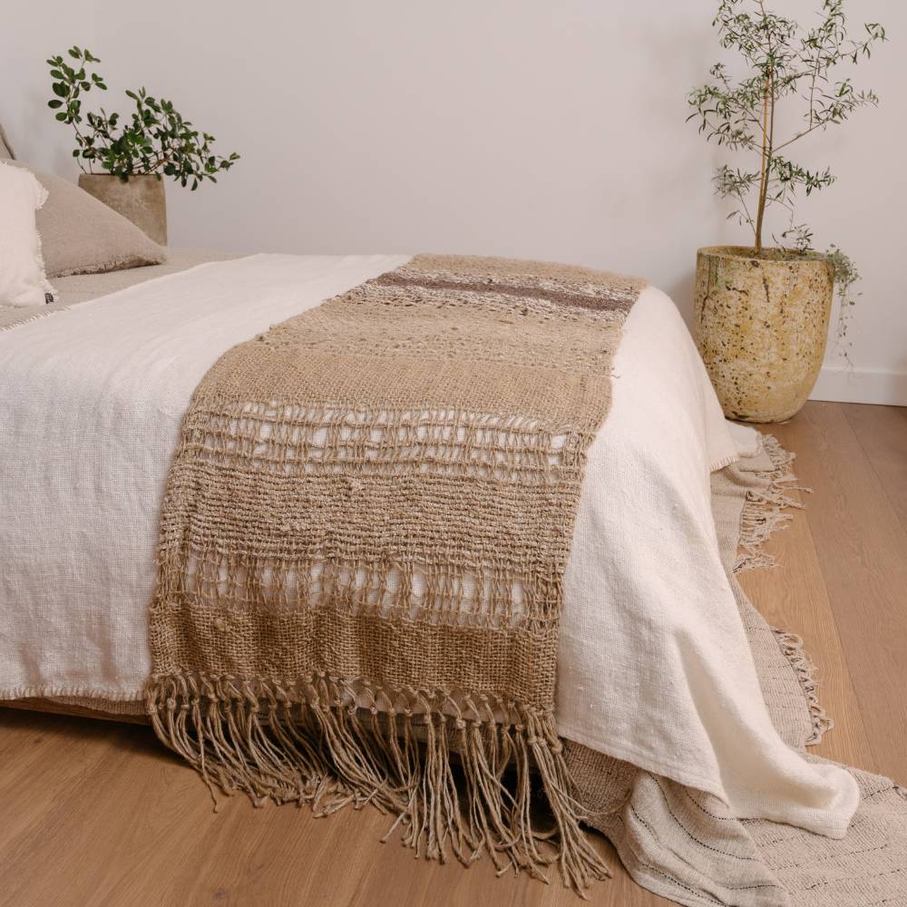 Textured blanket draped over a bed with a neutral color scheme, featuring a plant in the background.