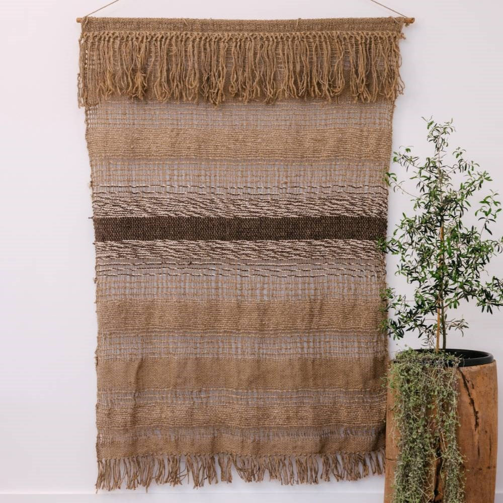 Handwoven natural jute wall hanging with fringes on a white wall next to a potted plant
