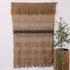 Handwoven natural jute wall hanging with fringes on a white wall next to a potted plant