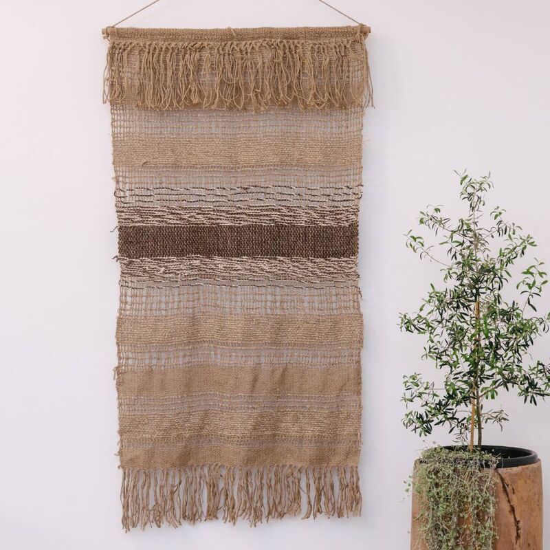 Handwoven natural jute wall hanging with fringes on a white wall next to a potted plant