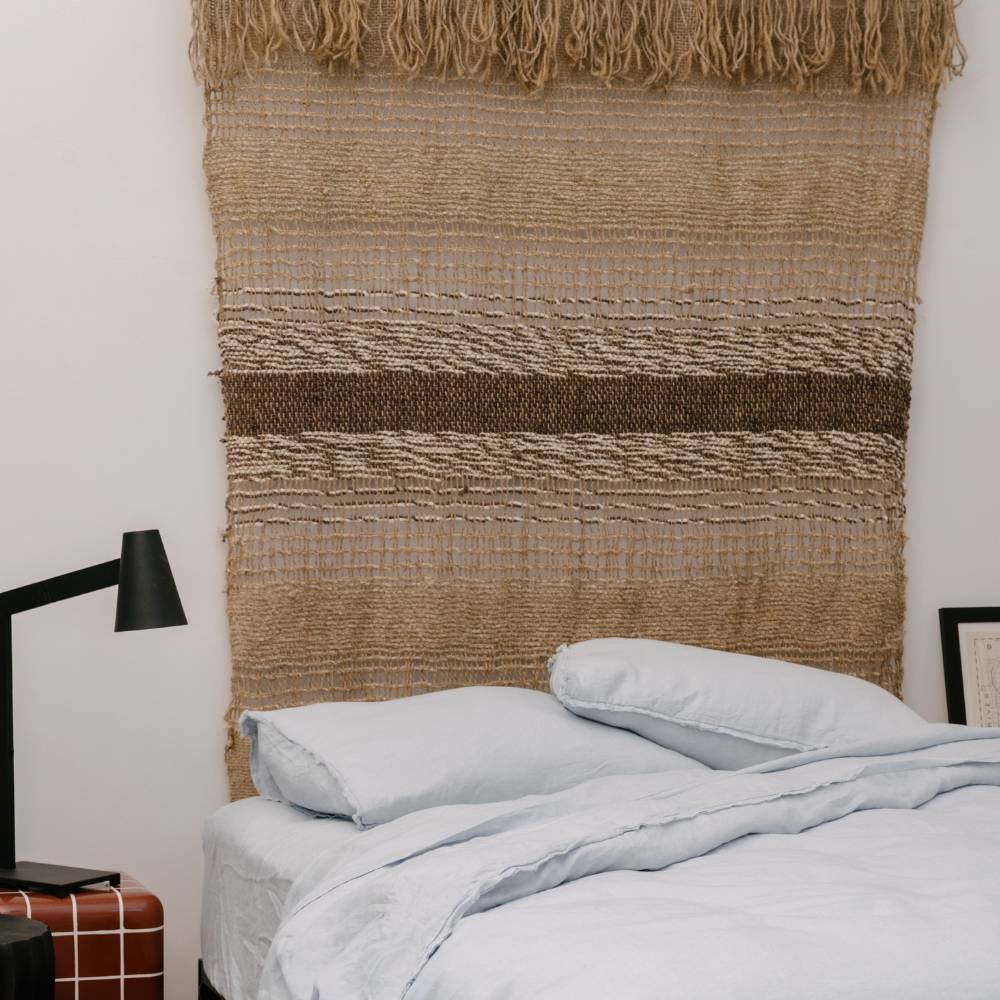 Handwoven natural jute wall hanging with fringes on a white wall next to a potted plant