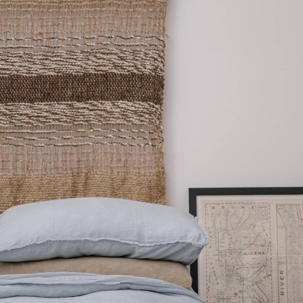 Handwoven natural jute wall hanging with fringes on a white wall next to a potted plant