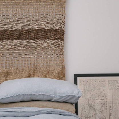 Handwoven natural jute wall hanging with fringes on a white wall next to a potted plant