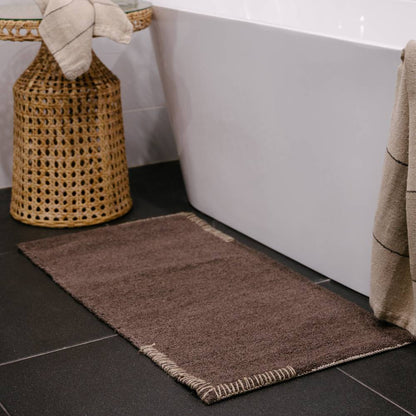 Dark brown bathmat and towel on a dark tiled bathroom floor with a white bathtub.
