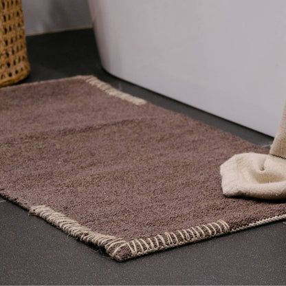 Dark brown bathmat and towel on a dark tiled bathroom floor with a white bathtub.
