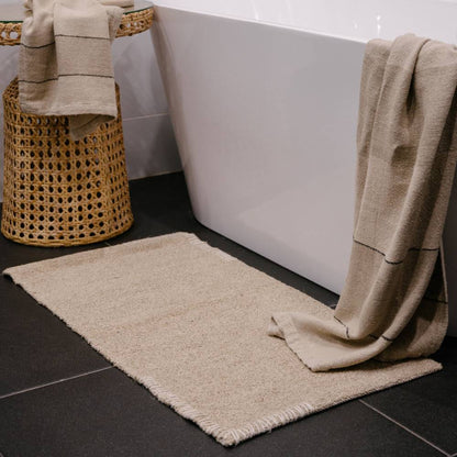 Beige bathmat and towel on a dark tiled bathroom floor with a white bathtub.