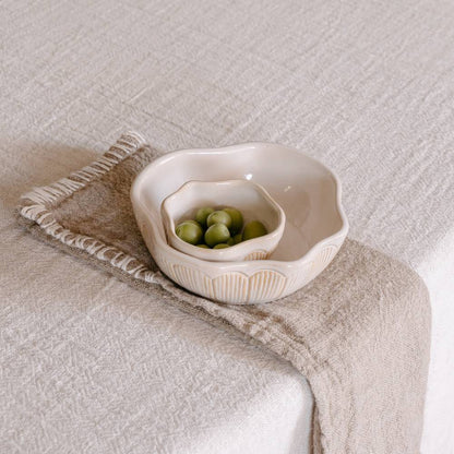 A textured beige linen table napkin with a decorative stitched edge is draped over a light tablecloth, holding two bowls.