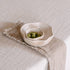 A textured beige linen table napkin with a decorative stitched edge is draped over a light tablecloth, holding two bowls.