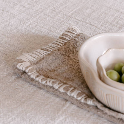 A textured beige linen table napkin with a decorative stitched edge is draped over a light tablecloth, holding two bowls.