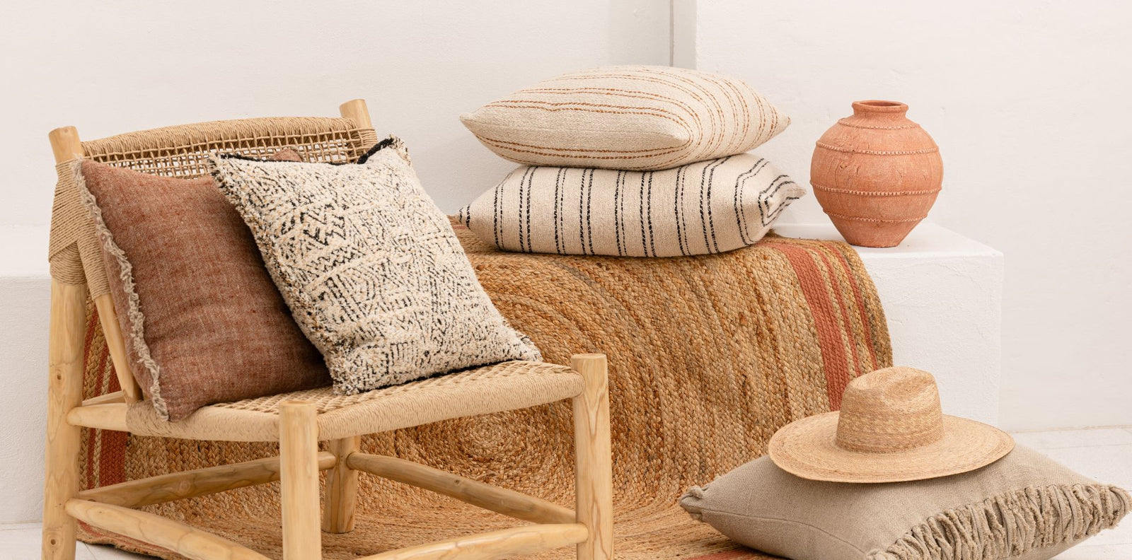 Woven chair with textured linen Eadie cushions and a hat, set against a white wall. Summer mediterranean feel.