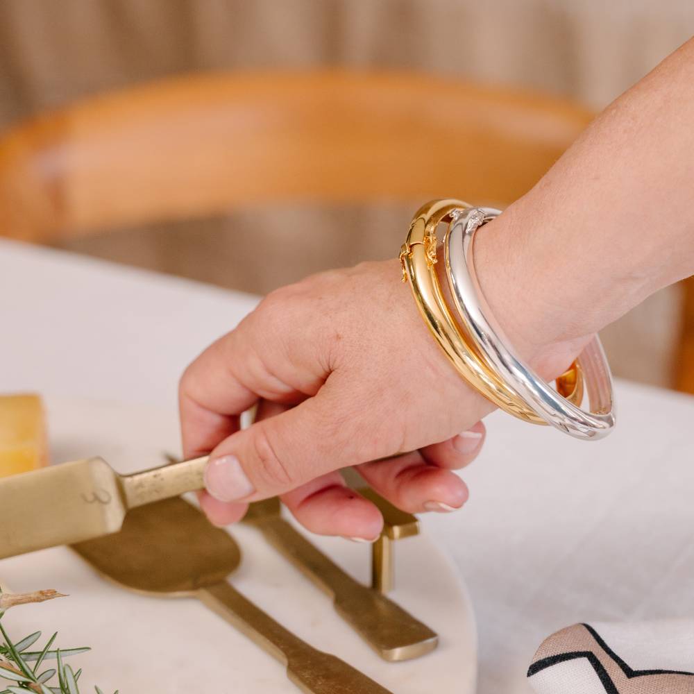 Hand holding a gold cheese knife with two bangles on a neutral background
